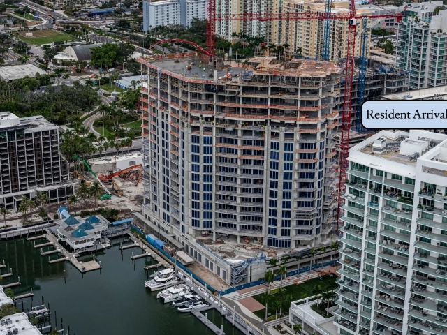 Drone photograph showing a multi-story concrete residential tower under construction along a marina. Two red tower cranes extend above the building, and workers are visible on the rooftop slab. Boat slips and docked vessels line the water&rsquo;s edge. Nearby are completed condominium buildings, palm trees, and city streets in a coastal urban setting.