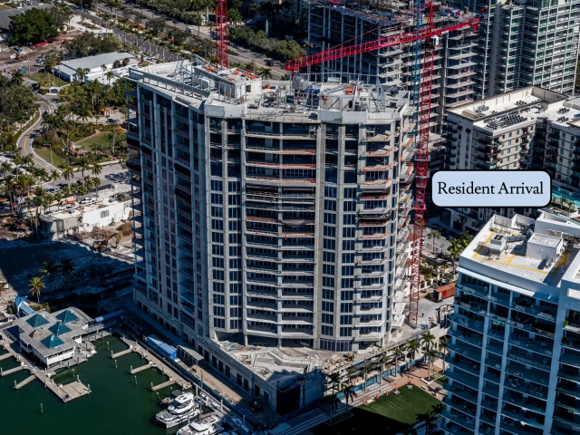 Curved high-rise residential tower under construction with red cranes by marina.