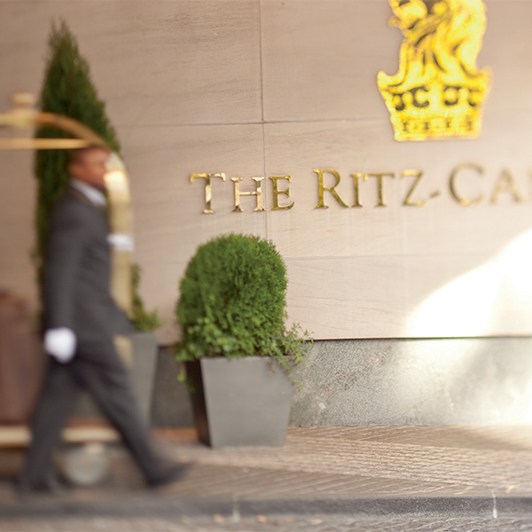 Uniformed staff member walking past The Ritz-Carlton entrance sign with luggage cart.