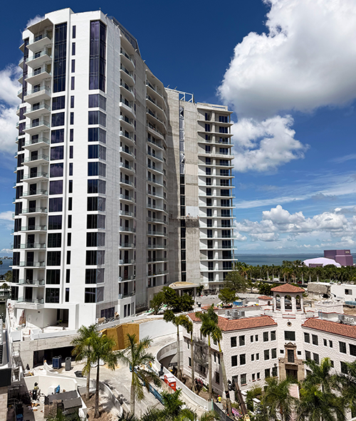 A luxury high-rise condominium tower under construction rises above Mediterranean-style buildings and palm trees along Florida's waterfront skyline.